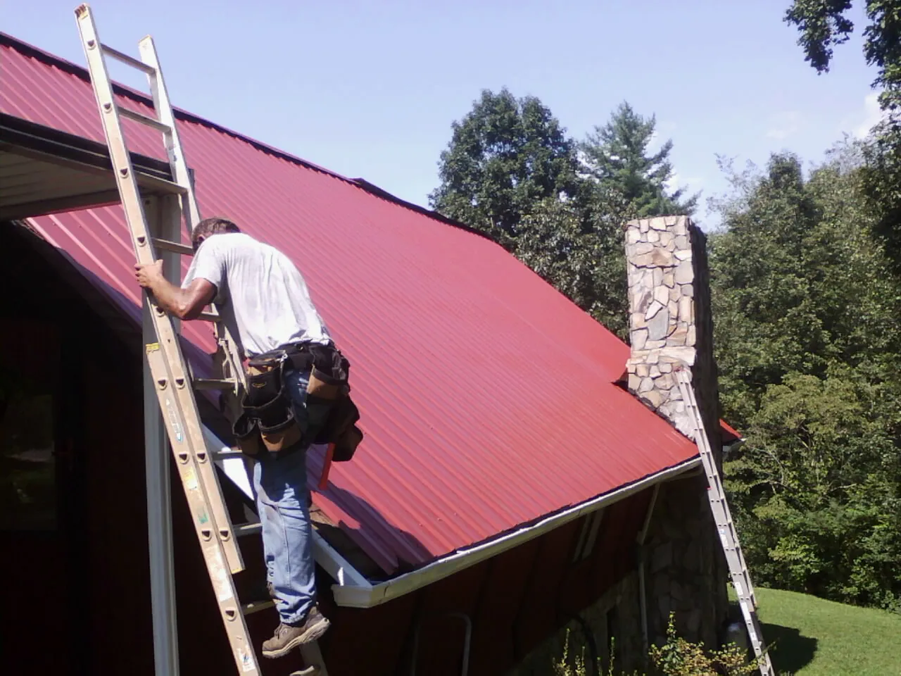 Roofer on a ladder beside a red metal roof and stone chimney — Soffit & Fascia Repair by Roofing Brothers in Oakton
